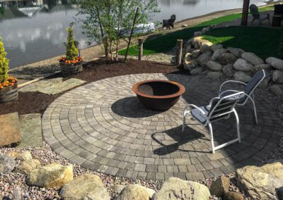 Circular stone patio with center firepit and chairs facing the lake